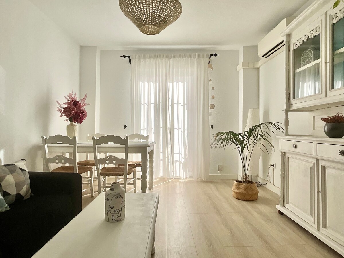 A bright living area features a light-colored sofa and a white coffee table. A dining table with four chairs is positioned near large glass doors, which are dressed in sheer white curtains. A decorative plant and a vase add a touch of elegance to the space.