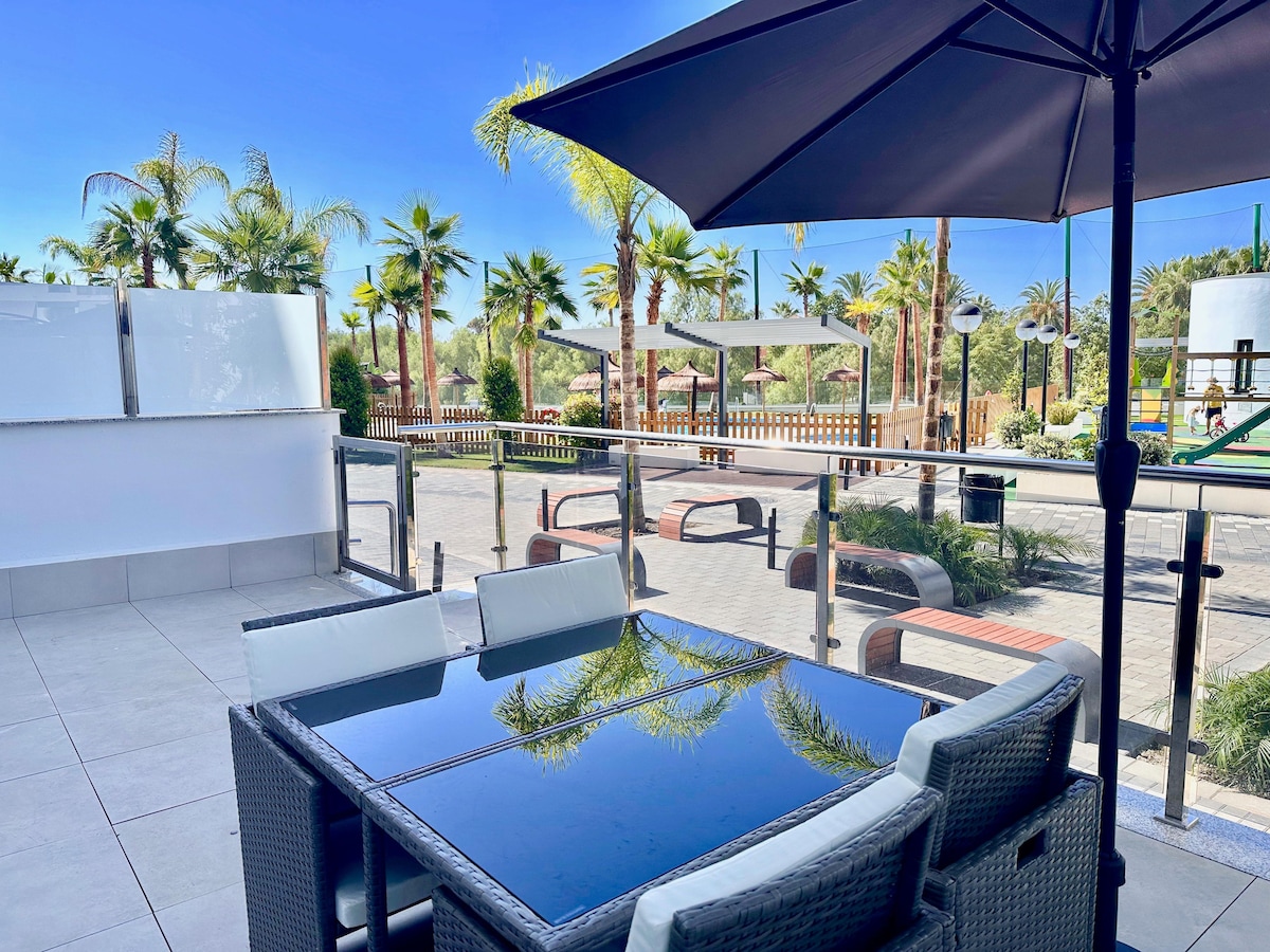 A terrace area features a glass table surrounded by comfortable chairs, shaded by a large umbrella. In the background, palm trees and loungers create a tropical atmosphere, while recreational spaces are visible. The layout promotes relaxation and social gatherings.