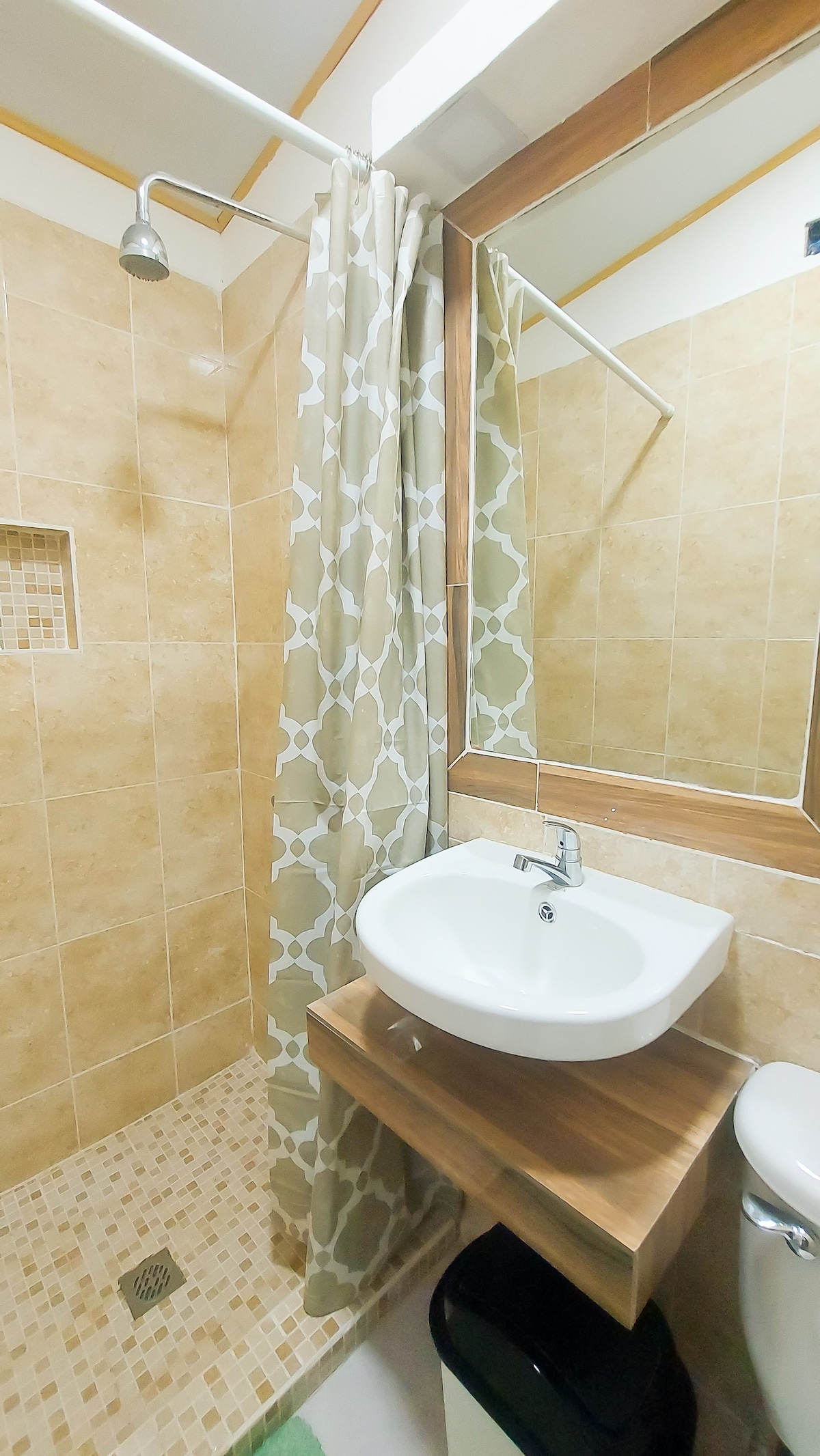A private bathroom is presented with beige tiled walls and floor. A glass shower is highlighted with a patterned curtain. A modern basin is mounted on a wooden shelf, with a mirror above enhancing the space. A black trash bin is positioned beside the toilet.