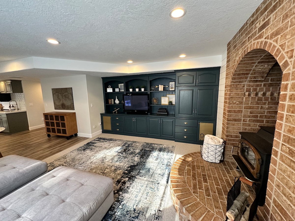 A spacious living area is highlighted by a large sectional sofa and a decorative area rug. A custom-built shelving unit in a deep blue tone is positioned against the wall, with a flat-screen TV and various decorative items displayed. An ornate brick fireplace adds a touch of warmth.