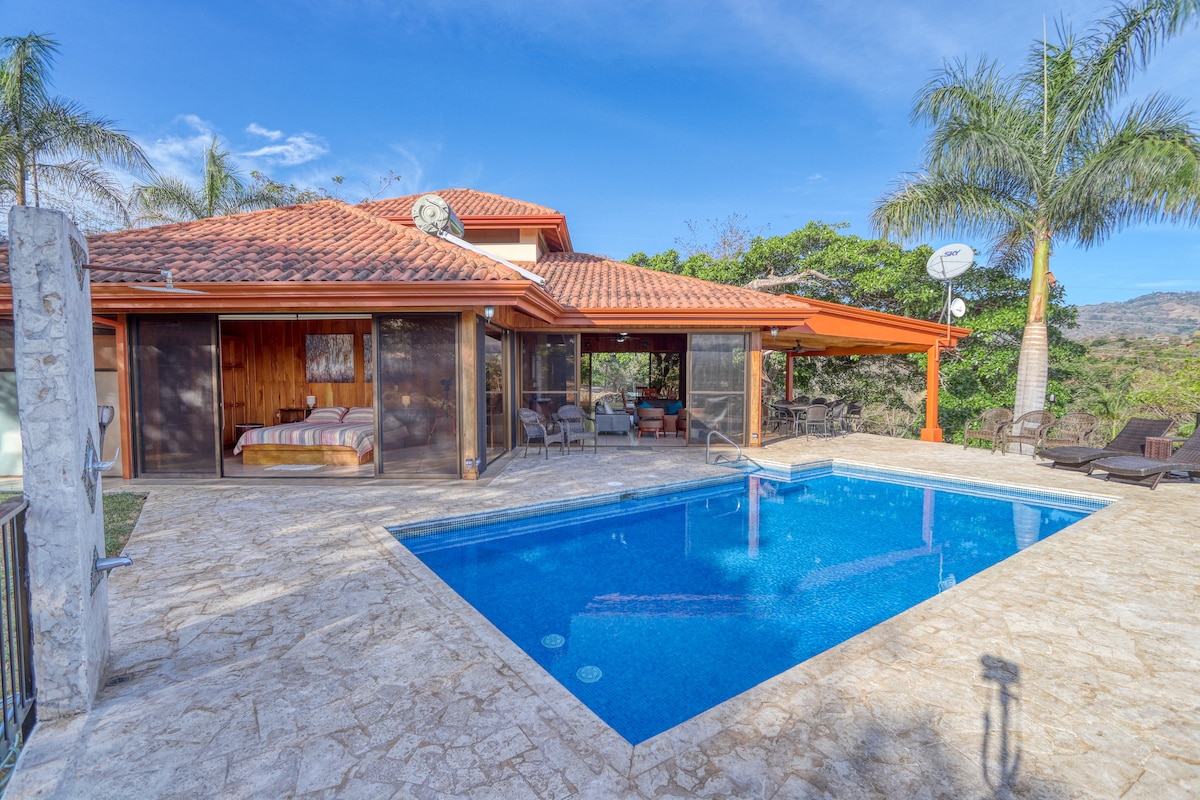 A spacious outdoor area features a crystal-clear pool, surrounded by a sunlit patio with lounge chairs and a dining set. Lush greenery is visible in the background, while the villa's inviting terrace provides shaded seating, enhancing the serene atmosphere.