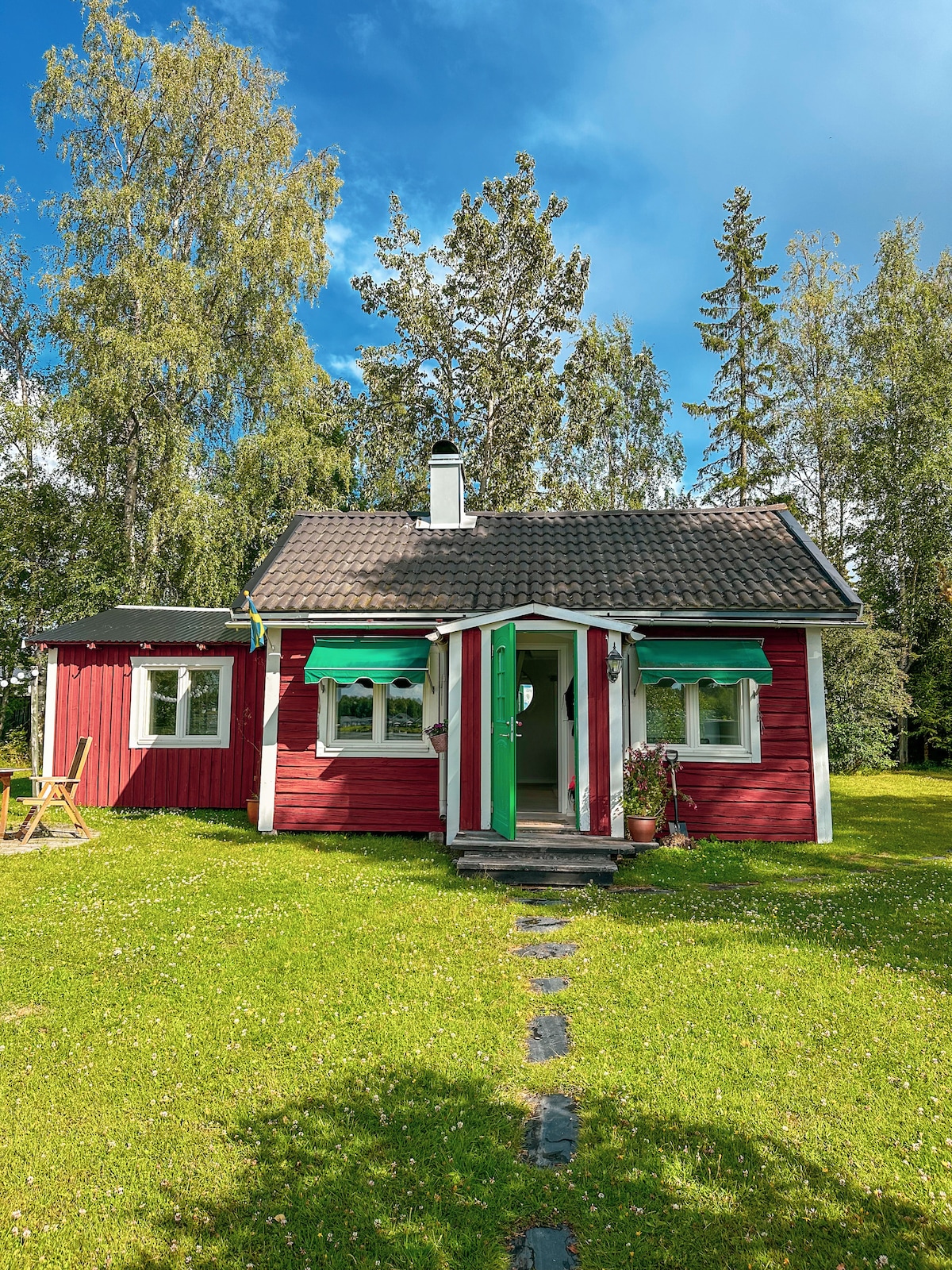 A charming red cottage is set amidst lush greenery, featuring a welcoming green door and decorative awnings. A small pathway leads to the entrance, complemented by a peaceful grassy area and surrounded by trees, providing a serene atmosphere.