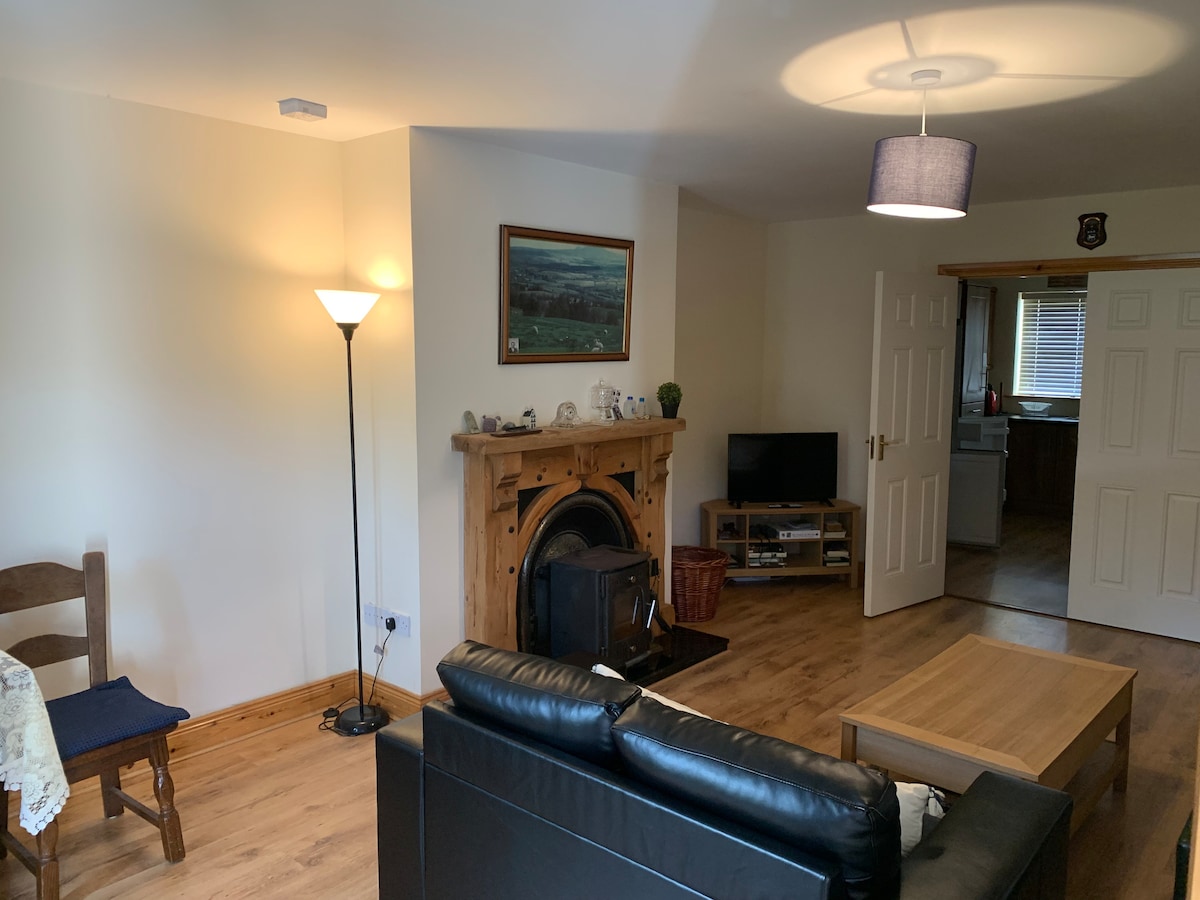 A cozy living space features a dark leather sofa positioned near a wooden coffee table. A lamp and a floor lamp provide soft lighting, while a decorative fireplace adds warmth. An artwork hangs on the wall, and a television is visible in the background.