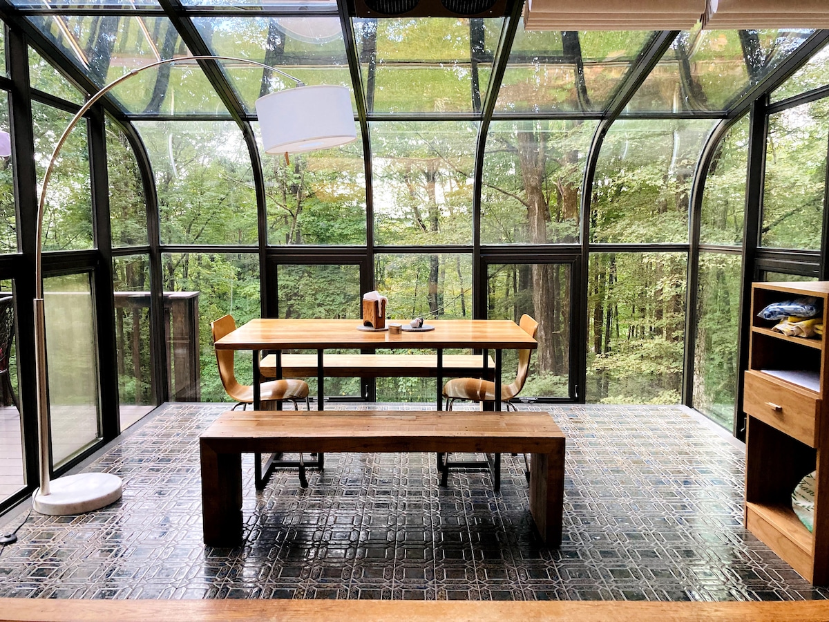 A spacious solarium features large glass walls, filled with natural light and offering views of the surrounding trees. A wooden table is centered in the room, accompanied by several chairs and a long bench. Warm wood tones are complemented by a patterned tile floor.