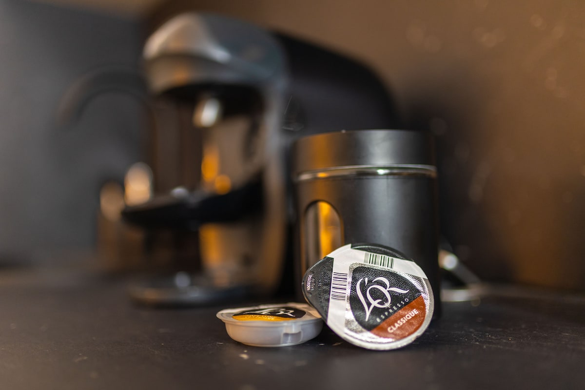 A compact coffee maker is positioned in the background, while a single coffee pod rests prominently in the foreground. The dark surfaces of the kitchen counter create a modern look, emphasizing the coffee-making setup.
