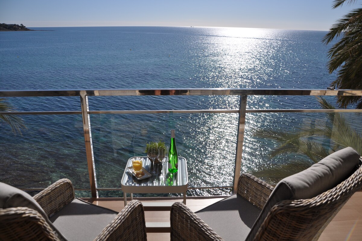 A sunlit terrace overlooks the tranquil sea, with gentle waves reflecting sunlight. Two comfortable rattan chairs are positioned around a small table, which holds bottles of sparkling water and a bowl of snacks. Lush greenery adds a touch of nature to the scene.