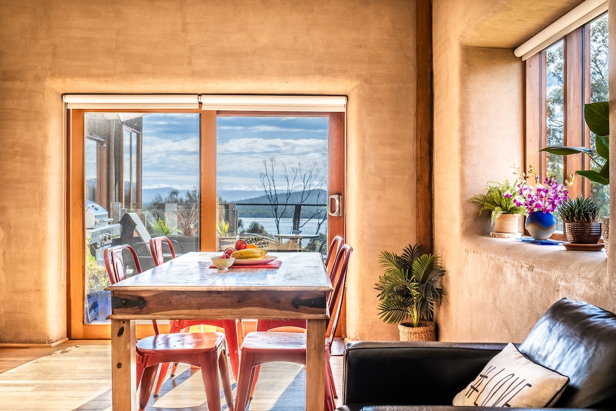 A dining area is presented with a wooden table accompanied by red chairs. Large glass doors provide a view of the water and distant landscape, while plants decorate the windowsills, enhancing the natural ambiance of the space.