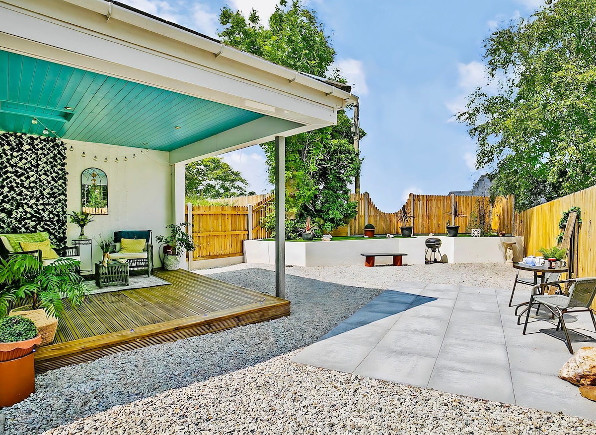 An outdoor area features a gravel surface bordered by a wooden fence. A covered porch with wooden flooring is furnished with comfortable seating. The yard has a patio space with smooth tiles, surrounded by greenery and decorative elements, providing a relaxing outdoor retreat.