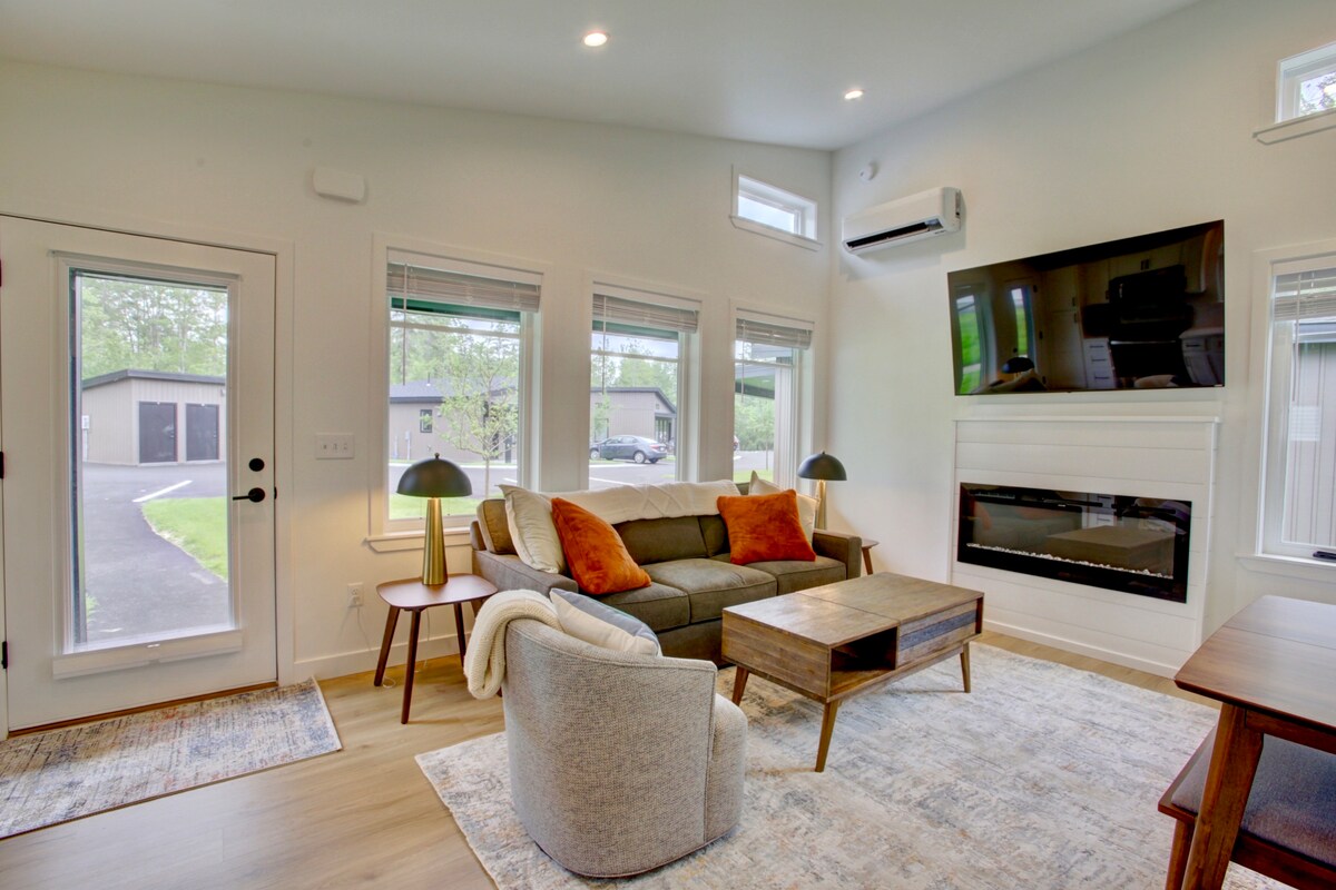 A modern living area is presented with natural light filtering through large windows. A comfortable couch is accented with throw pillows, facing a wooden coffee table and an electric fireplace. An air conditioning unit is mounted above, with side tables and a rug enhancing the space.