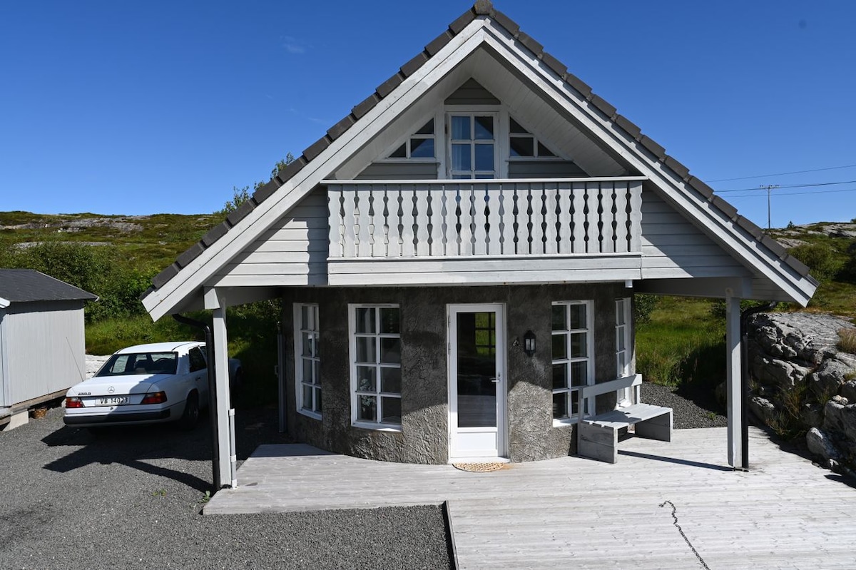 The exterior view of a two-story chalet showcases a wooden balcony and large windows allowing for natural light. A gravel pathway leads to the entrance, and a parked vehicle is visible to the side. Lush greenery surrounds the property, emphasizing its tranquil setting.