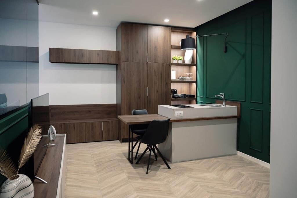 A modern kitchenette is featured with sleek cabinetry in warm wood tones. A minimalist dining table and chairs occupy the space, while a wall-mounted light adds illumination. The overall design incorporates green accents and is complemented by light-colored herringbone flooring.