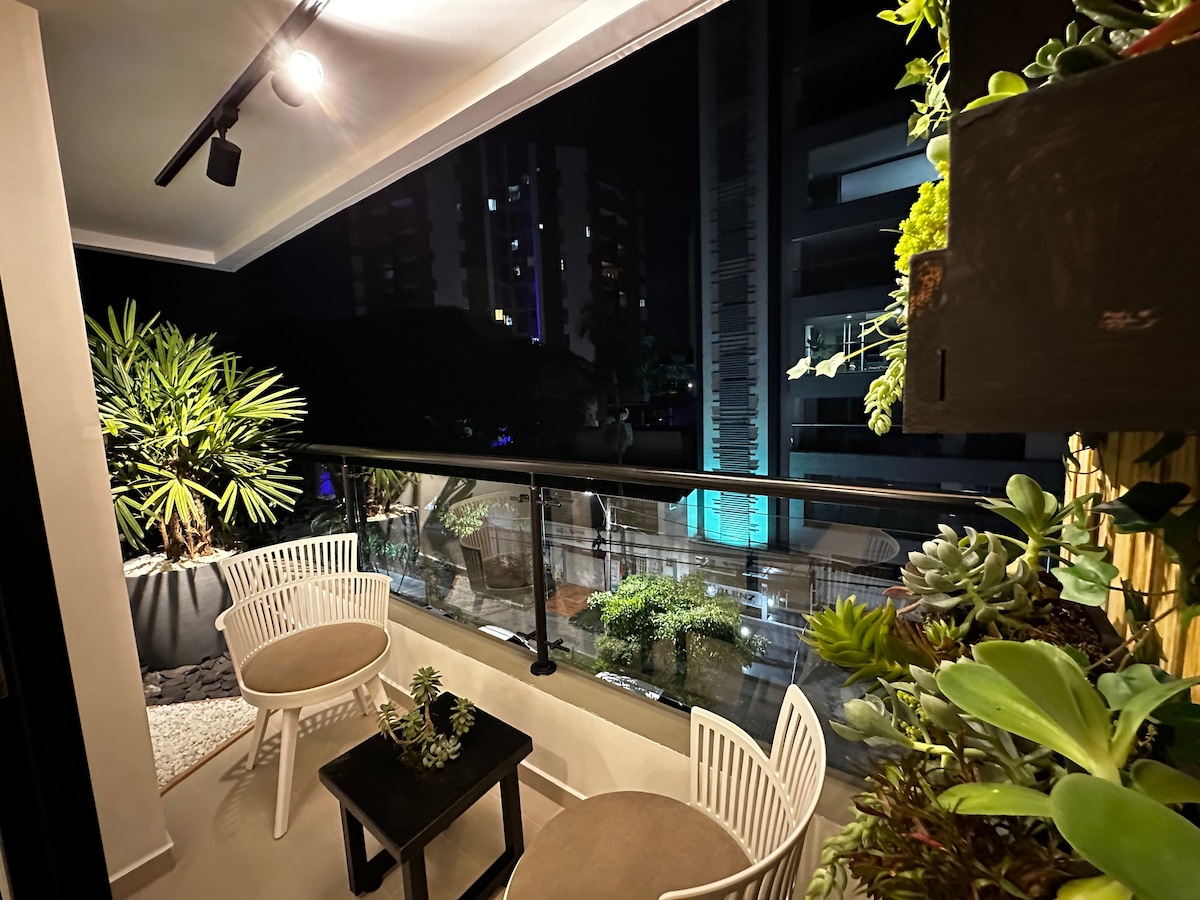 A balcony area features two modern chairs and a small black table, surrounded by potted plants. The glass railing offers views of the surrounding buildings and greenery, with soft lighting illuminating the space at night.