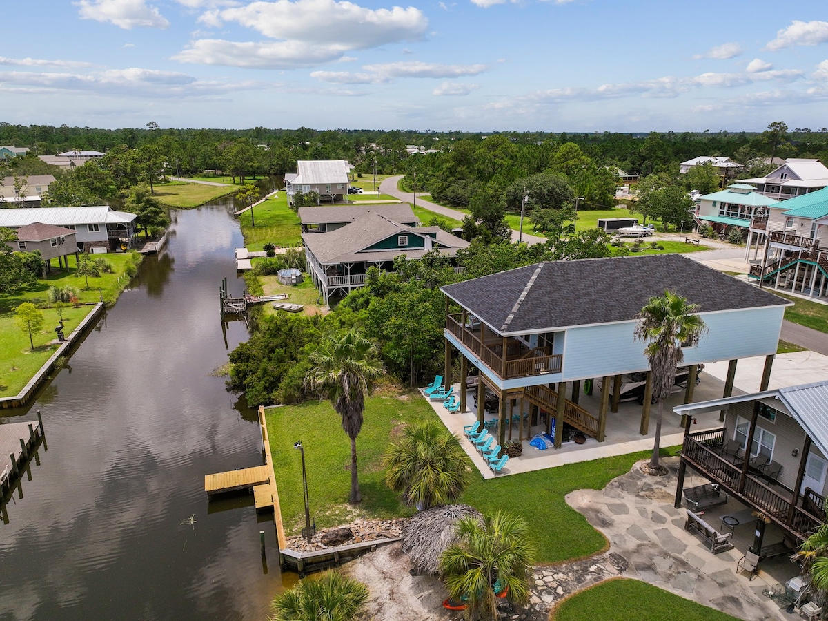 An aerial view showcases a modern holiday home adjacent to a deep-water canal. Lush greenery surrounds the property, and multiple chairs can be seen along the canal's edge for relaxation. Nearby structures are visible, indicating a peaceful community setting.