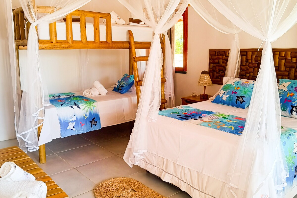 A cozy sleeping area features a spacious double bed alongside a comfortable bunk bed. Light bed linens and colorful decorative pillows add to the decor. A small bedside lamp illuminates the space, while a woven rug and wooden elements bring warmth to the room.