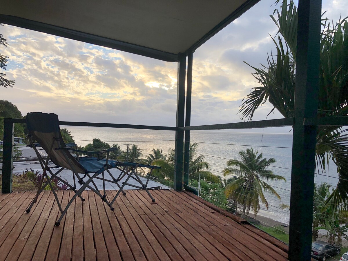 A covered balcony offers a panoramic view of Savusavu Bay and the ocean. Two folding chairs are positioned on a wooden deck, highlighting the spacious layout. Lush palm trees and a cloudy sky create a serene backdrop, inviting relaxation and outdoor enjoyment.