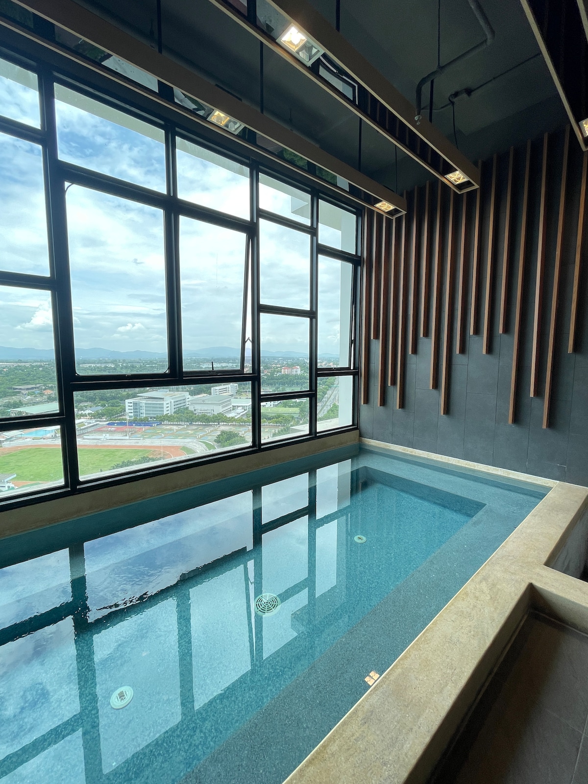 An indoor swimming pool is showcased beside large windows, offering a panoramic view of the surrounding area. The pool features a sleek design with clear blue water reflecting natural light. Wooden accents on the wall enhance the contemporary aesthetic.