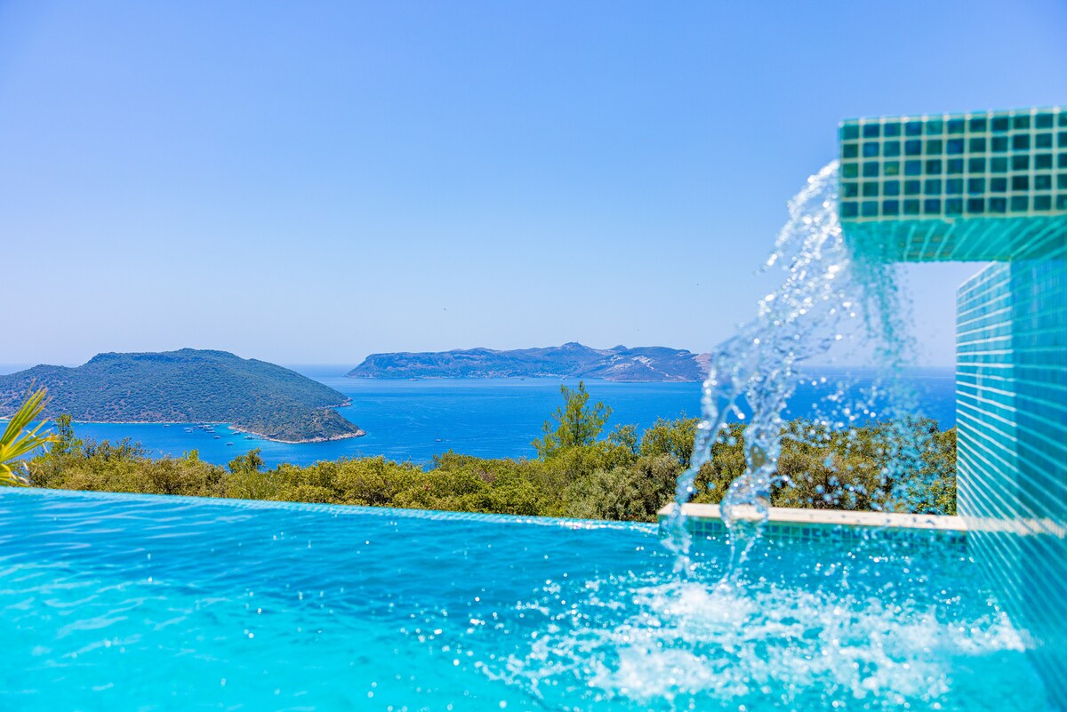 An infinity pool with clear blue water extends towards the horizon, where it meets the tranquil sea. A waterfall feature cascades gently into the pool, surrounded by lush greenery and scenic views of the islands in the distance.