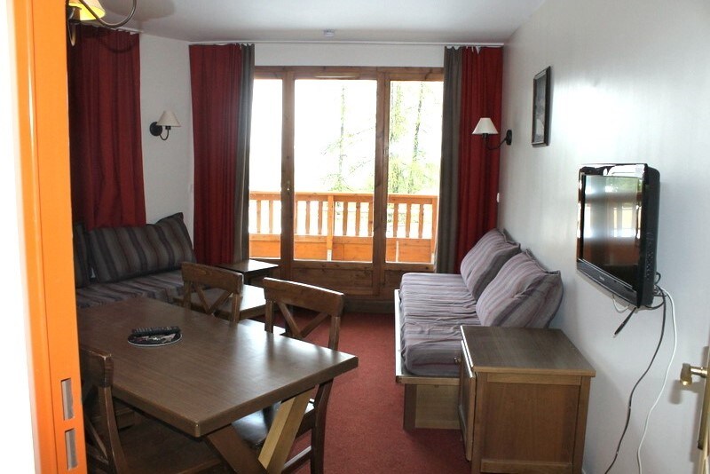 A comfortable living area is presented with a wooden dining table and a television mounted on the wall. Two seating areas, including a striped sofa and a couple of cushioned benches, face large glass doors that open to a balcony with wooden railings.