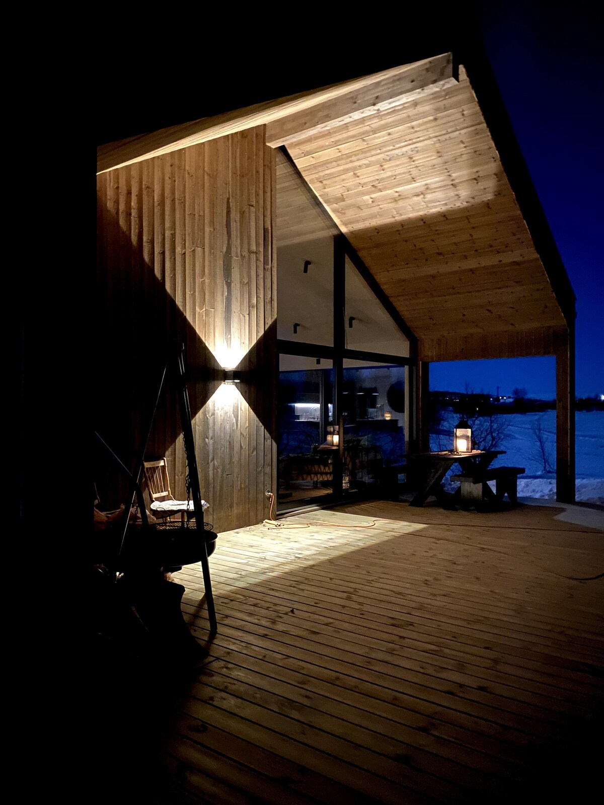 The exterior of a wooden cabin features a spacious deck illuminated by warm lighting. A sloped roof creates a striking architectural detail, while large glass panels offer views of the surrounding landscape. Rustic seating elements are positioned alongside the entrance.