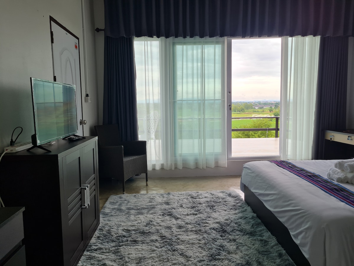 A spacious bedroom features a large bed covered with soft linens and a colorful throw. A flat-screen television is positioned on a cabinet, while a comfortable chair and a sliding glass door invite natural light and provide access to the balcony overlooking the surrounding fields.