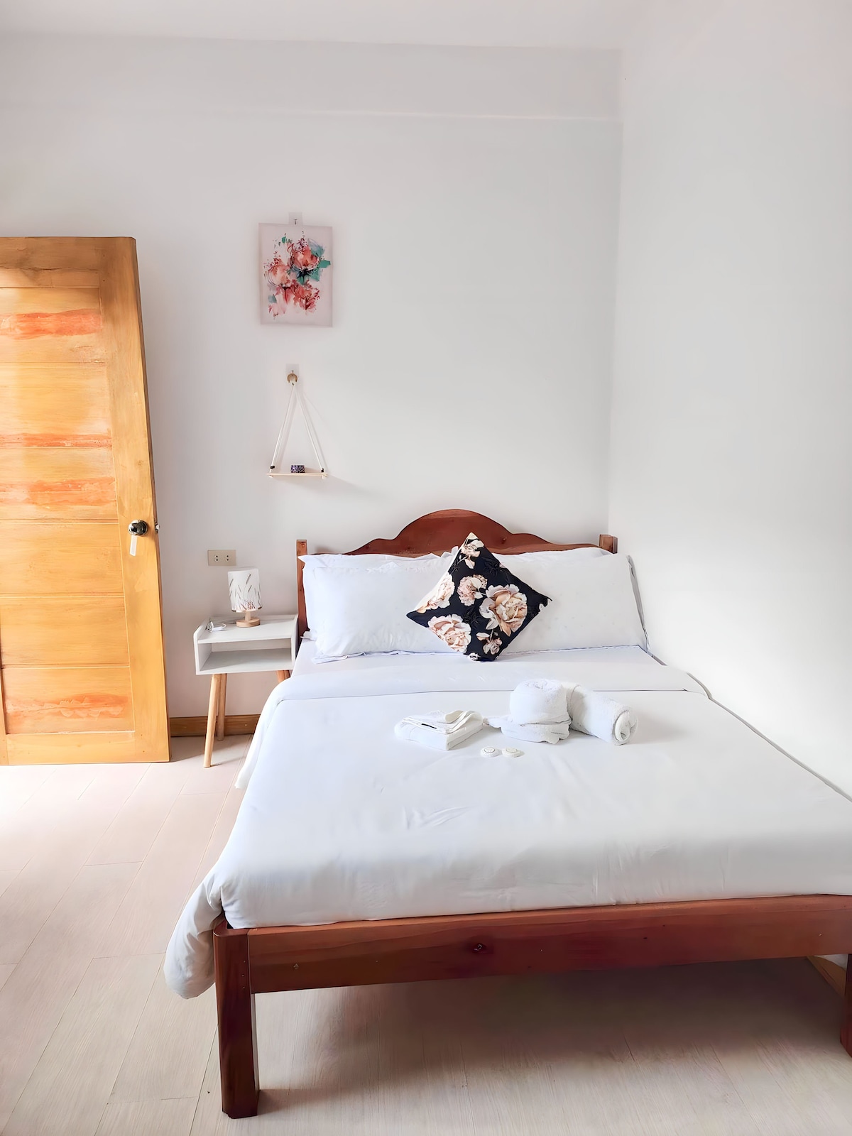A well-lit bedroom features a wooden bed with white linens and a decorative floral pillow. A small nightstand is positioned beside the bed, and neatly arranged towels are placed on the bed, complementing the room's minimalist design. A wooden door and a wall art piece add to the ambiance.