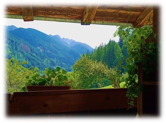 A scenic view of mountains and lush greenery is framed by a wooden railing. The landscape showcases rolling hills and towering trees under a clear sky, inviting a sense of tranquility and connection to nature.