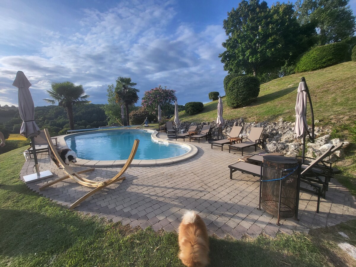 A serene outdoor area features a curved swimming pool surrounded by lounge chairs and umbrellas. Lush greenery and trees provide a natural backdrop. A pet is seen on the patio, adding a sense of tranquility to the setting.