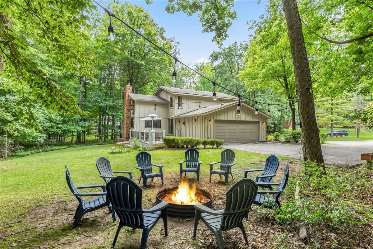An outdoor fire pit area features a circular arrangement of spacious, dark blue chairs around a central fire ring. String lights provide soft illumination above, with a lush wooded backdrop visible behind a light-colored house and garage.