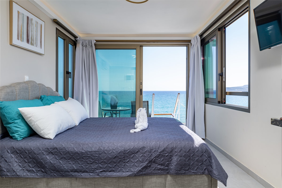 A spacious sleeping area is highlighted by a large bed dressed in soft linens and vibrant pillows. Floor-to-ceiling windows offer unobstructed views of the sea, while natural light creates a calming environment. A wall-mounted television is visible in the corner.