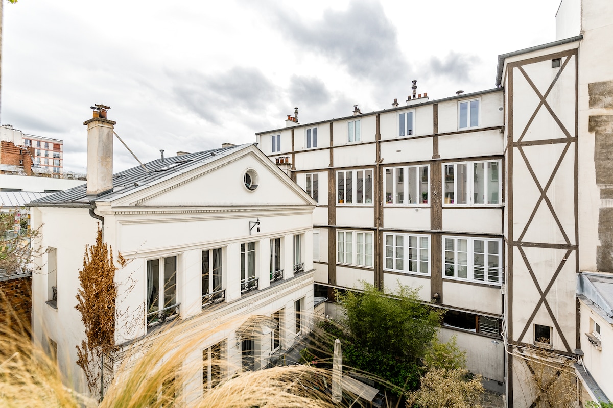 The view showcases a charming courtyard surrounded by classic Parisian architecture. A white building with large windows is framed by greenery, while neighboring structures exhibit traditional and modern styles. Soft natural light filters through the space, contributing to a serene atmosphere.