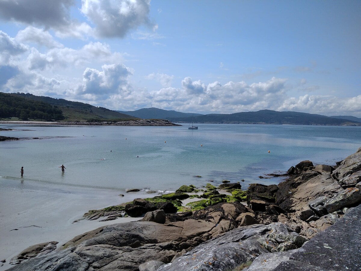 A serene coastal scene features calm waters meeting a rocky shoreline. Gentle ripples reflect soft cloud patterns overhead. Two individuals can be seen enjoying the water, while hints of green seaweed are visible on the rocks, adding to the natural beauty.