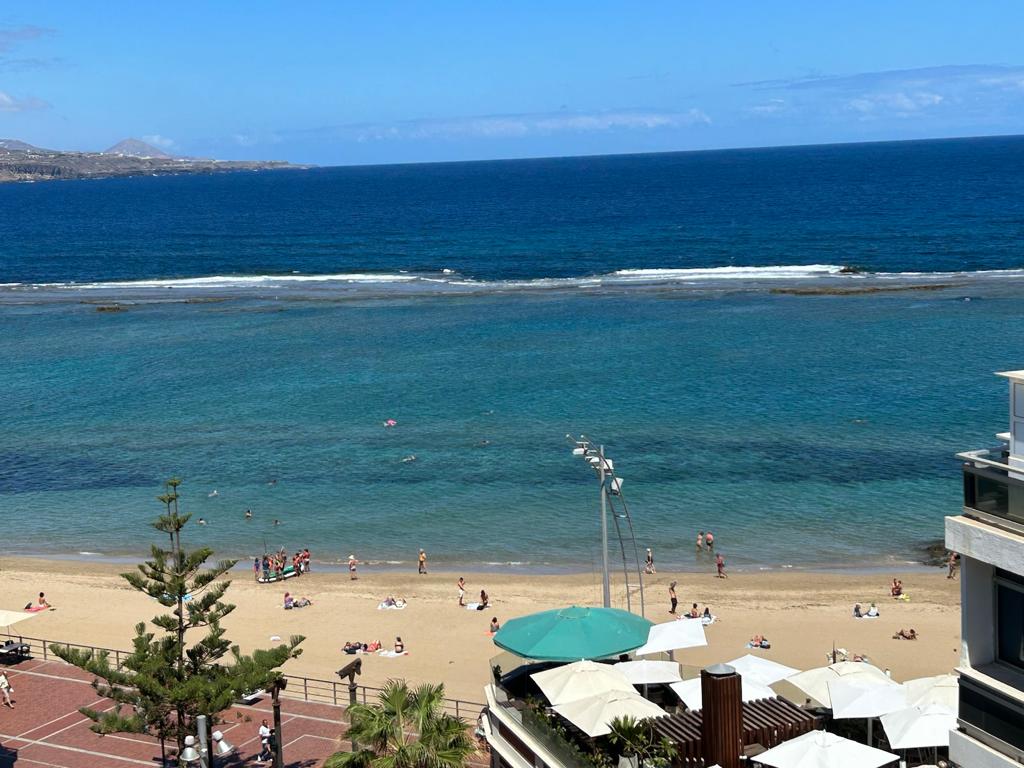 A coastal view features a sandy beach adjacent to a calm blue ocean. Small groups of beachgoers are visible enjoying the sun, while colorful umbrellas are stationed on the beach. The shoreline is lined with gentle waves, creating a serene atmosphere.