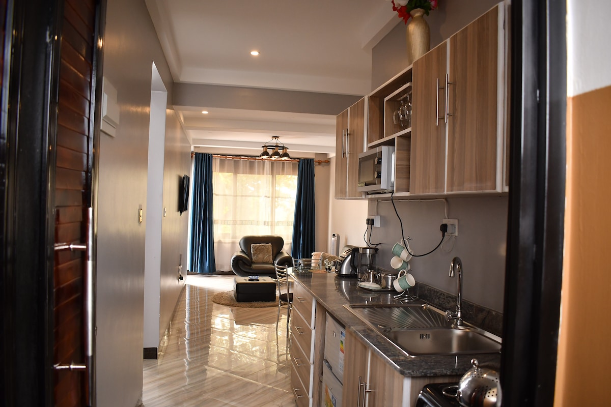 A modern kitchen area features wooden cabinetry and a polished countertop. Appliances such as a kettle and microwave are neatly arranged. Natural light fills the adjacent living space, where a comfortable chair and decorative elements are visible, enhancing the welcoming environment.