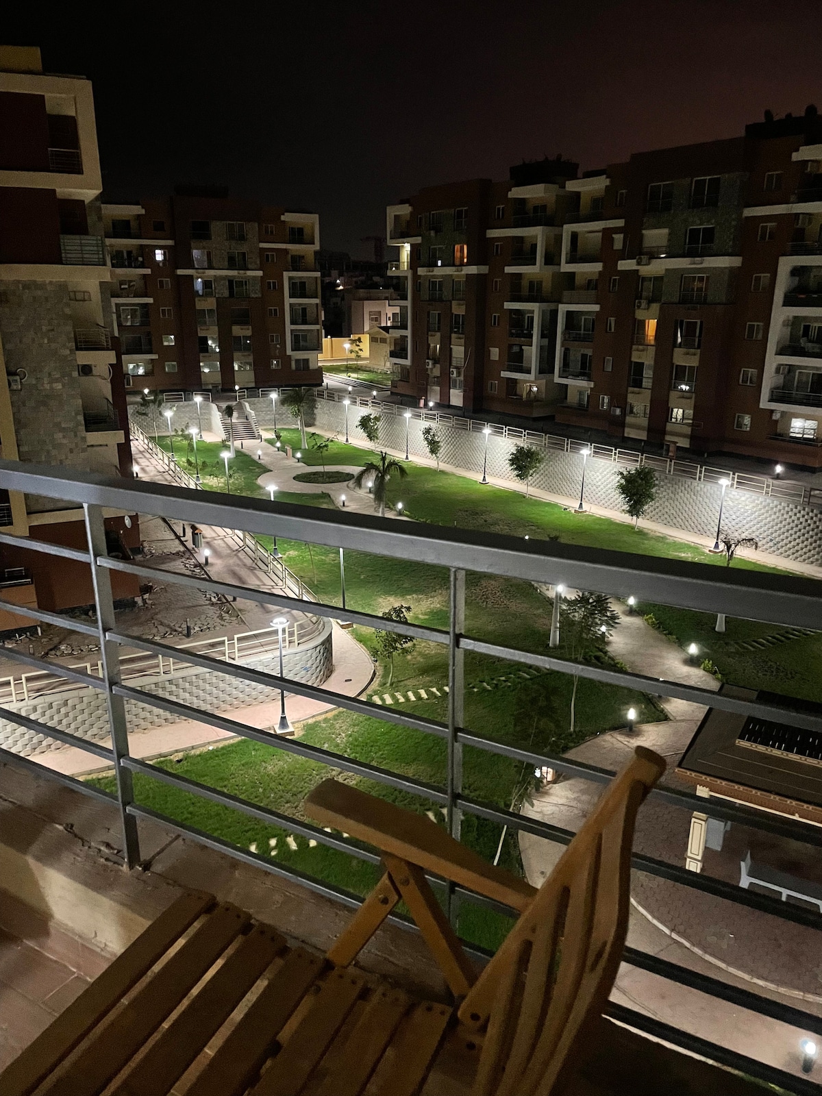 A nighttime view from a balcony reveals a well-lit garden area below, featuring pathways and greenery. Surrounding buildings display a modern architectural style, while a wooden chair is positioned on the balcony, providing a space to relax and enjoy the scenery.