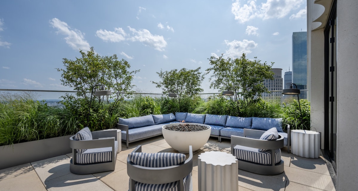 A spacious rooftop terrace features a circular fire pit surrounded by a blue seating area with cushions, framed by lush greenery. Two striped armchairs and a small round table add to the inviting ambiance, with the skyline visible in the background under a clear sky.