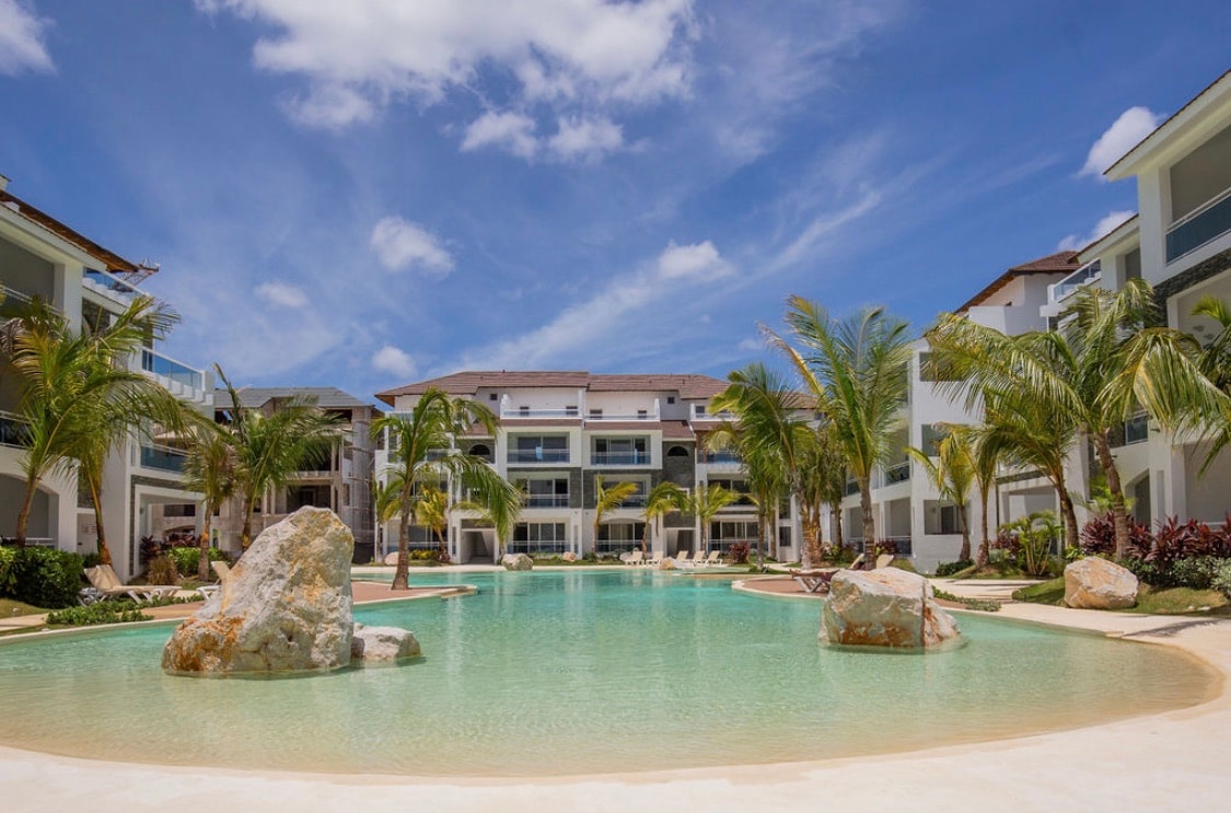 A serene pool area is surrounded by palm trees and lush landscaping, with large boulders incorporated into the design. Several lounge chairs are positioned around the water's edge, inviting relaxation under clear blue skies.