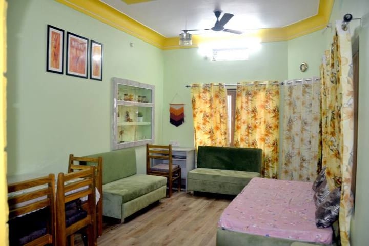 A well-lit living area features soft green walls and a stylish yellow ceiling. Two green sofas and a pink bed are arranged on the polished wooden floor, complemented by bright floral curtains. Framed artwork adorns the wall above a glass display cabinet.
