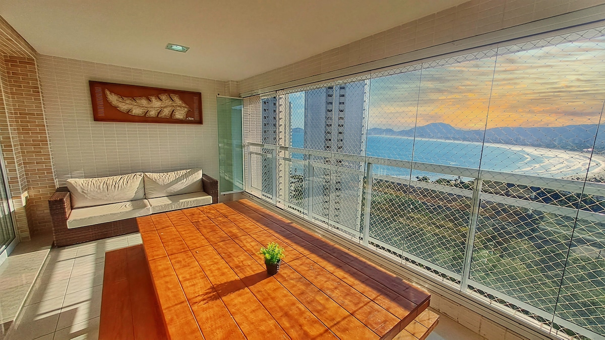 A spacious balcony features a wooden table complemented by a small potted plant. Comfortable seating is arranged nearby, with large glass windows providing an unobstructed view of the sea and surrounding landscape.