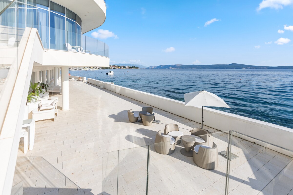A spacious terrace area features modern outdoor seating arrangements, including loungers and tables, overlooking the calm sea. An umbrella provides shade, while large windows of the adjacent building reflect the sunlight. The horizon offers a serene view of distant land under a clear blue sky.