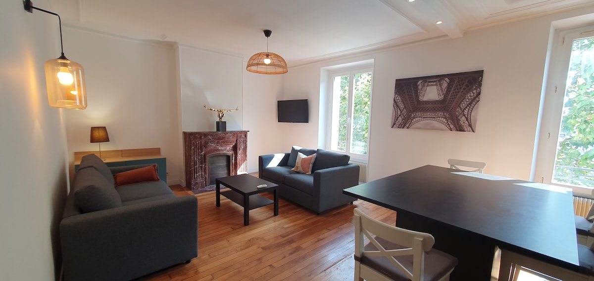 A spacious living area features two comfortable sofas and a coffee table, illuminated by natural light from large windows. A flat-screen TV is mounted on the wall, while a decorative fireplace adds character. A dining table and chairs are positioned nearby for meal gatherings.