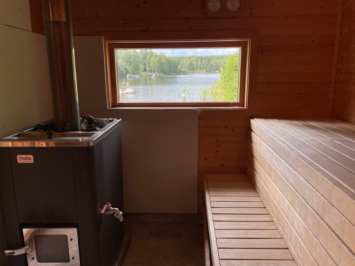 A wooden sauna interior features a sleek black stove and a spacious wooden bench. A large window provides a view of a tranquil lake surrounded by greenery, allowing natural light to fill the space.