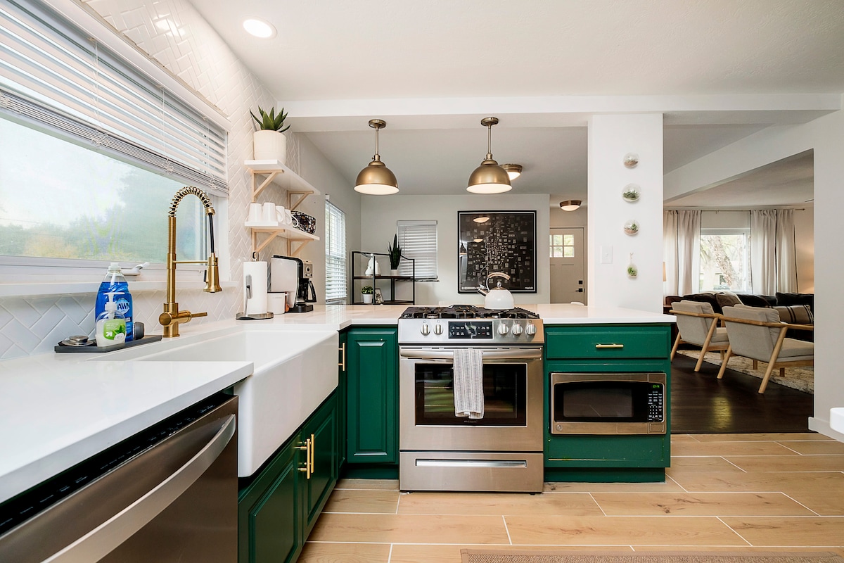 The kitchen features modern green cabinetry and a quartz countertop. A stainless steel gas range is positioned beside a built-in microwave. Stylish pendant lights hang above, illuminating the space. A large window allows natural light to enter, enhancing the open layout connecting to the living area.
