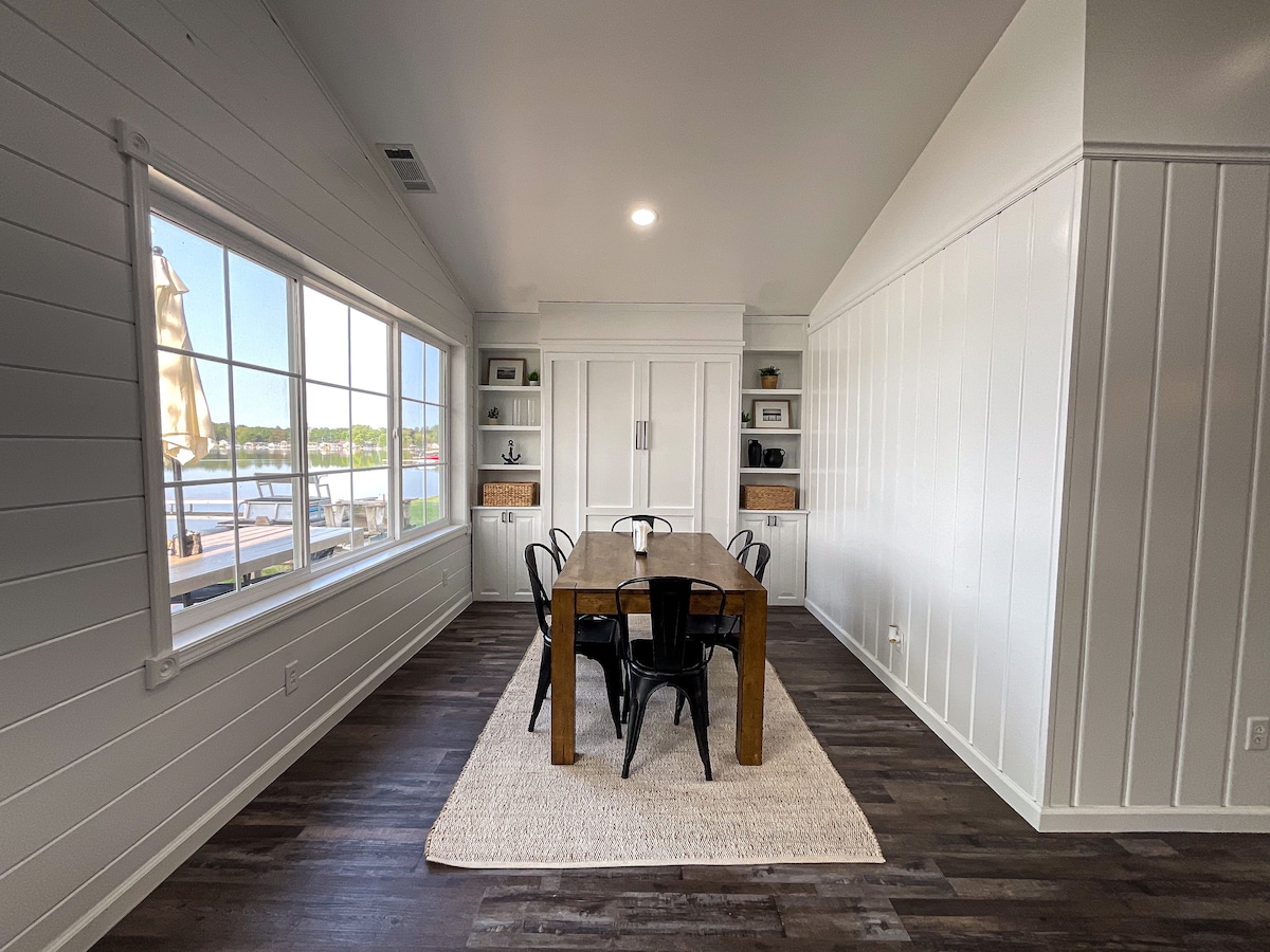 A bright dining room is furnished with a wooden table surrounded by six black chairs. A large window allows natural light to fill the space, offering views of the lake. The light-colored walls are complemented by an area rug under the table, enhancing the room's cozy feel.