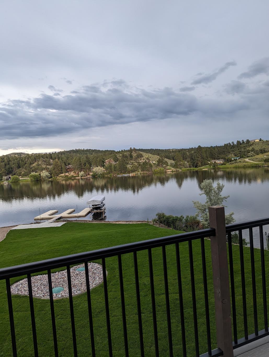 A serene view of Hauser Lake is captured from the balcony, showcasing calm waters reflecting the cloudy sky. The lush green lawn leads down to a dock where a boat is moored. Nearby, a landscaped area features pebbles and greenery.