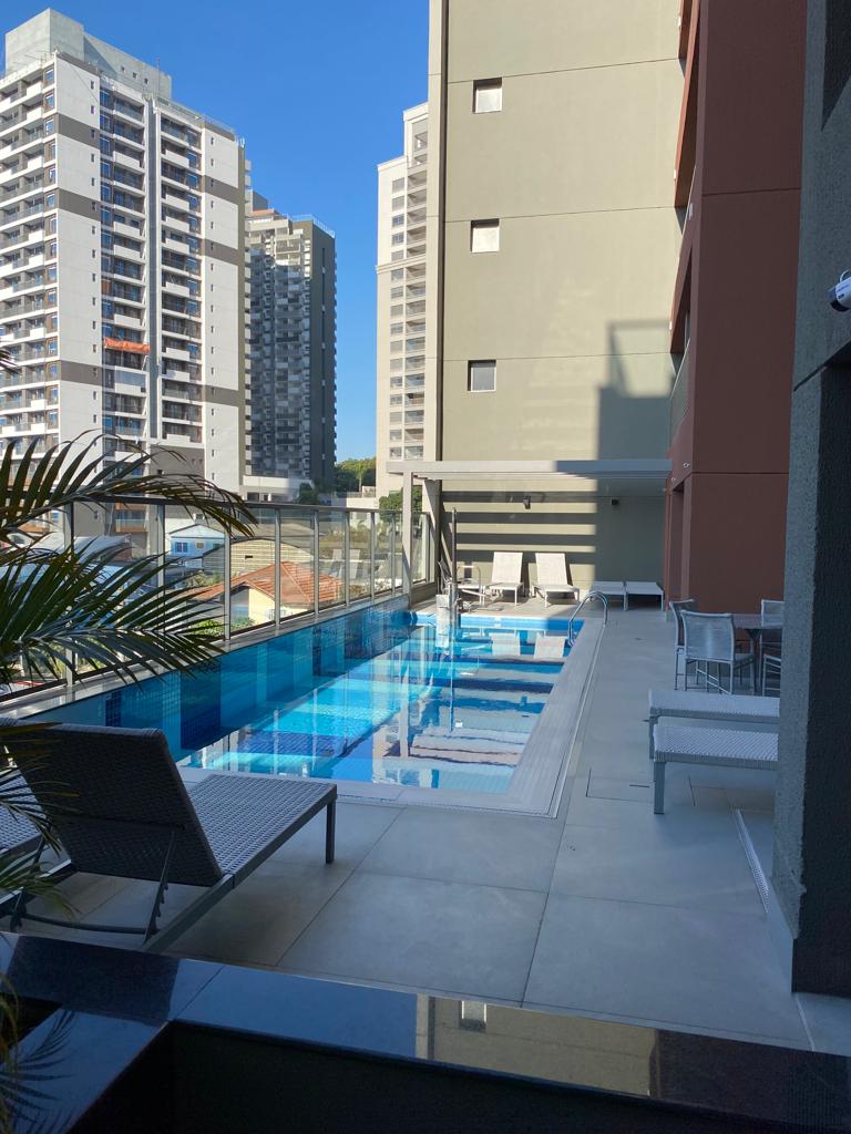 A modern swimming pool area is showcased, bordered by sleek lounge chairs. Sunlight reflects off the water, creating a vibrant blue hue. Surrounding high-rise buildings are visible in the background, highlighting the urban setting.