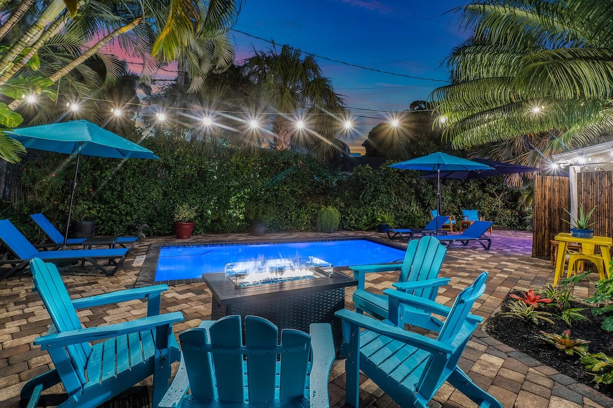 An inviting outdoor space features a heated pool, surrounded by lush greenery and palm trees. Blue lounge chairs and umbrellas are positioned nearby. A fire pit sits at the center, providing a cozy gathering spot, illuminated by string lights overhead as evening approaches.