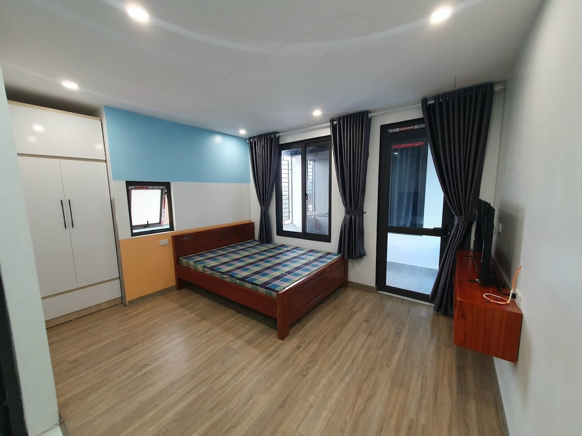 A well-lit bedroom features a spacious bed with a patterned blanket, accompanied by a wooden wardrobe and a minimalist television unit. The large windows with dark curtains allow natural light to enter, creating a serene atmosphere throughout the room.