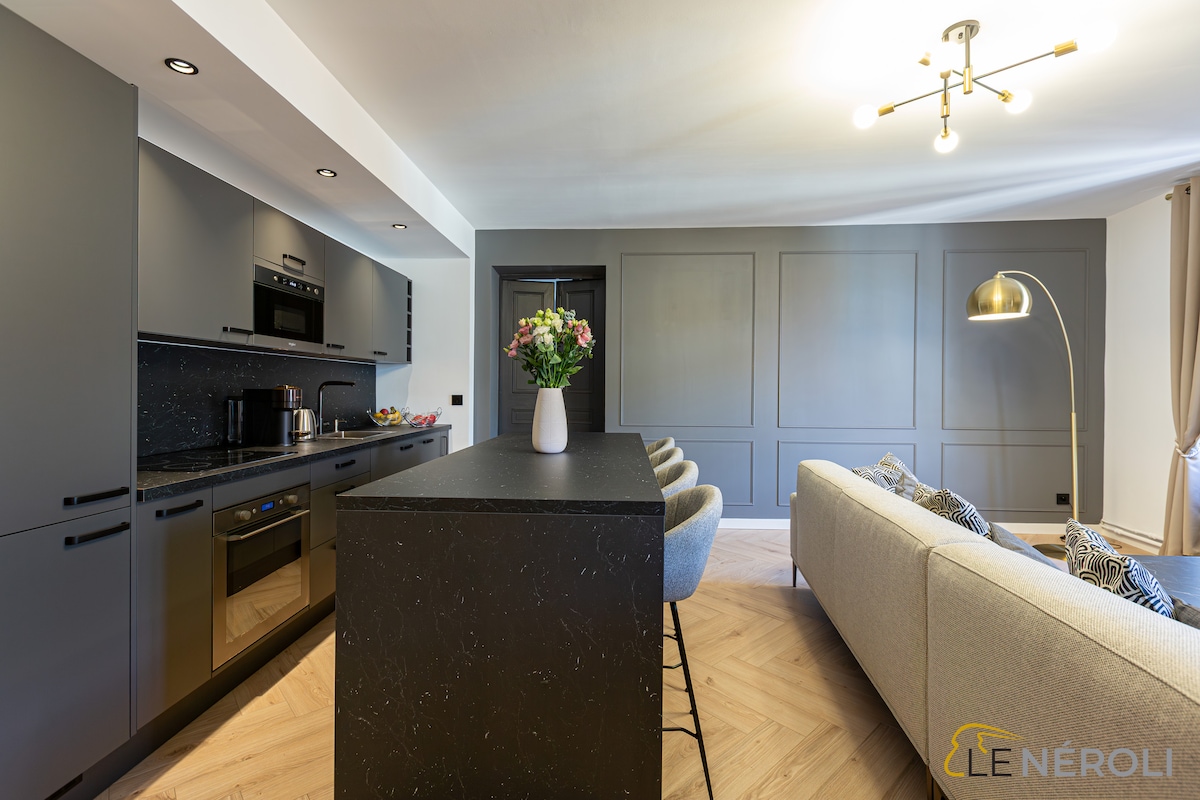 A modern kitchen area is highlighted by sleek cabinetry and an island with seating. A gray sofa is positioned nearby, complementing the minimalist design. Natural light illuminates the space through a nearby window, enhancing the contemporary feel.