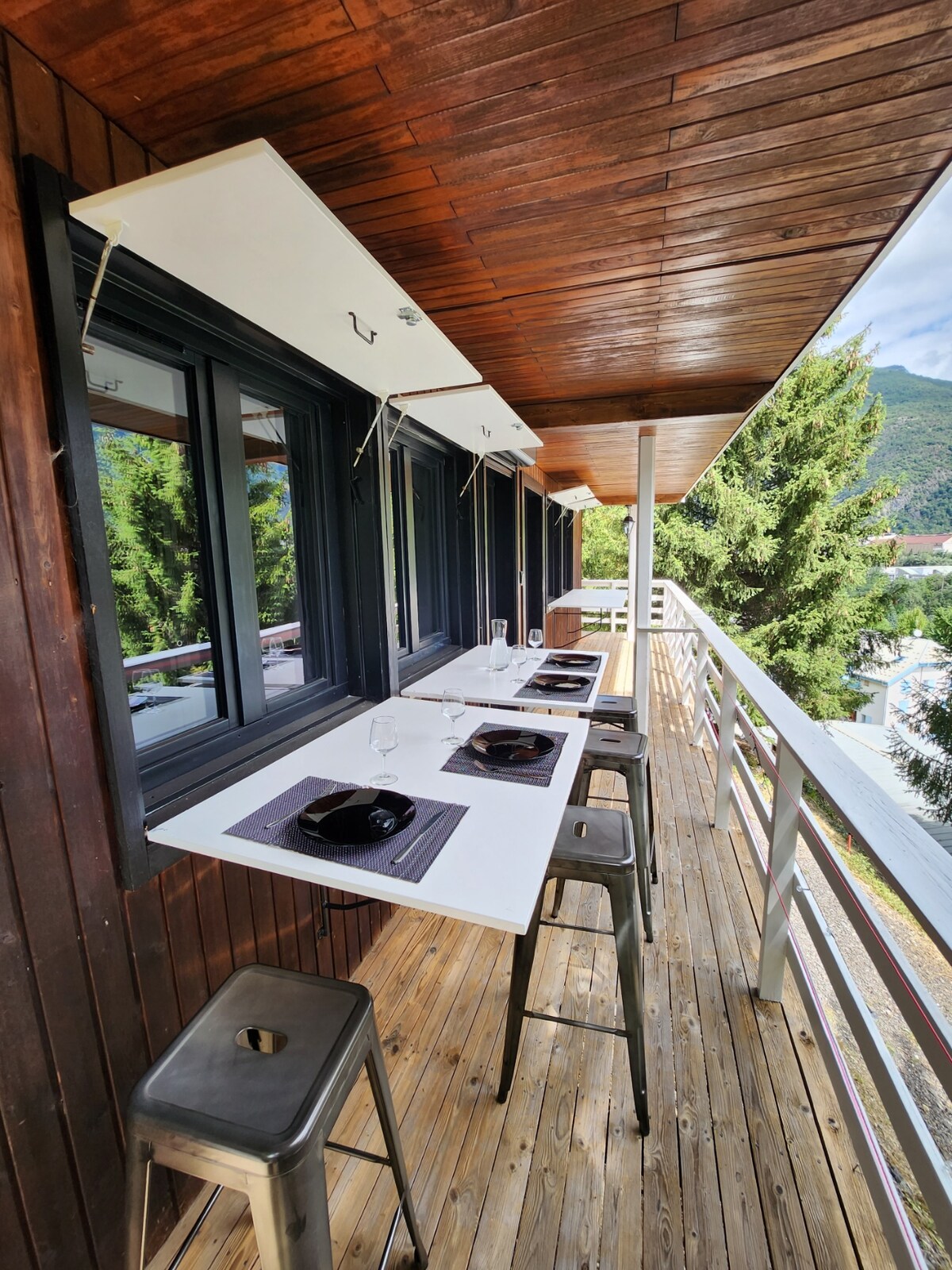 An outdoor terrace features a long, white table connected to the wall, with sleek metal stools positioned nearby. Place settings with black plates and cutlery are neatly arranged, providing a comfortable space for dining with views of the surrounding greenery.