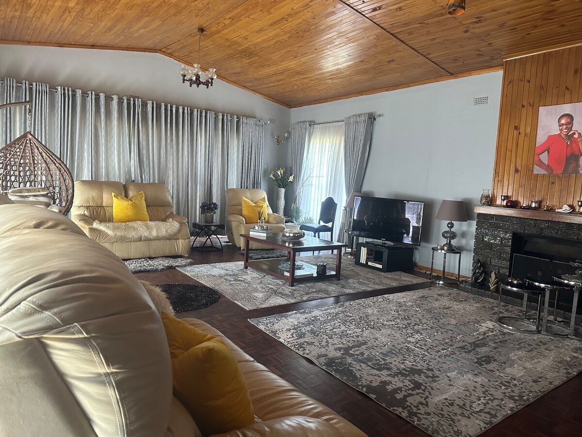 A spacious living area features high wooden ceilings and large windows allowing natural light to fill the room. Comfortable seating is arranged around a coffee table, with decorative cushions enhancing the space. A television and fireplace are positioned against a textured wall.